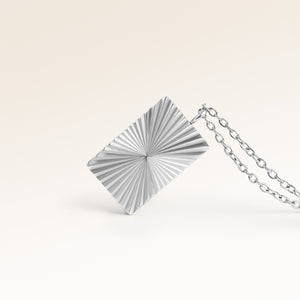 Närbild av minimalistiskt fyrkantigt berlock i silver med räfflad solstråle-yta och plats för gravyr baktill